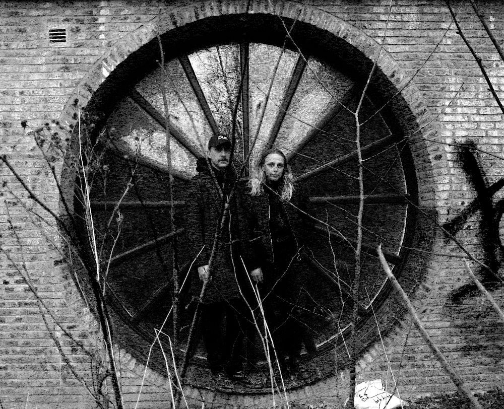 B&W photo of artists before a large circular metal window in brick. Svartvitt foto av två personer framför ett stort runt metallfönster i tegel.