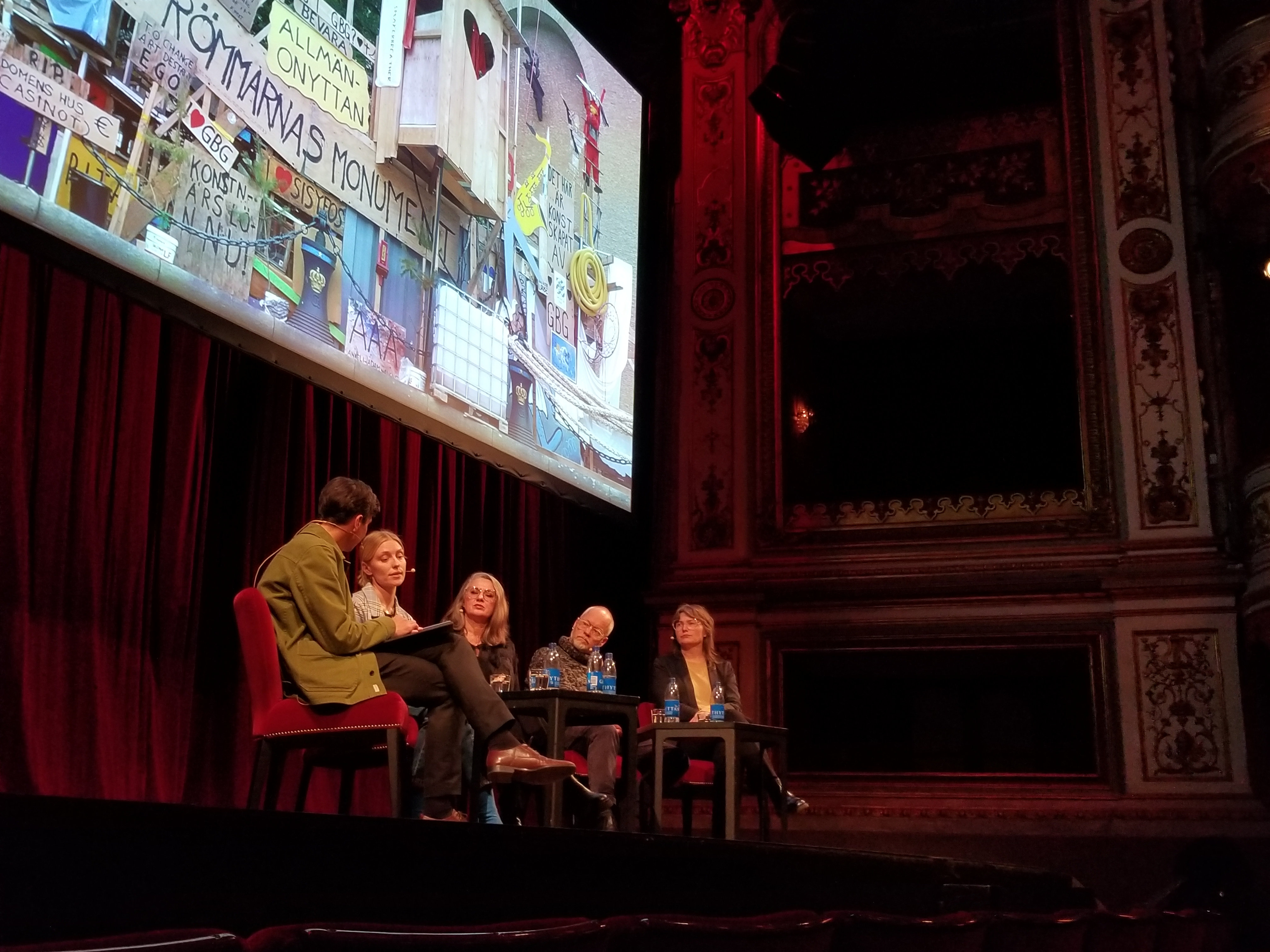 Stage panel discussing a large projected image of a chaotic wooden structure with signs. Panel diskuterar en projektion av ett komplext trämonument.