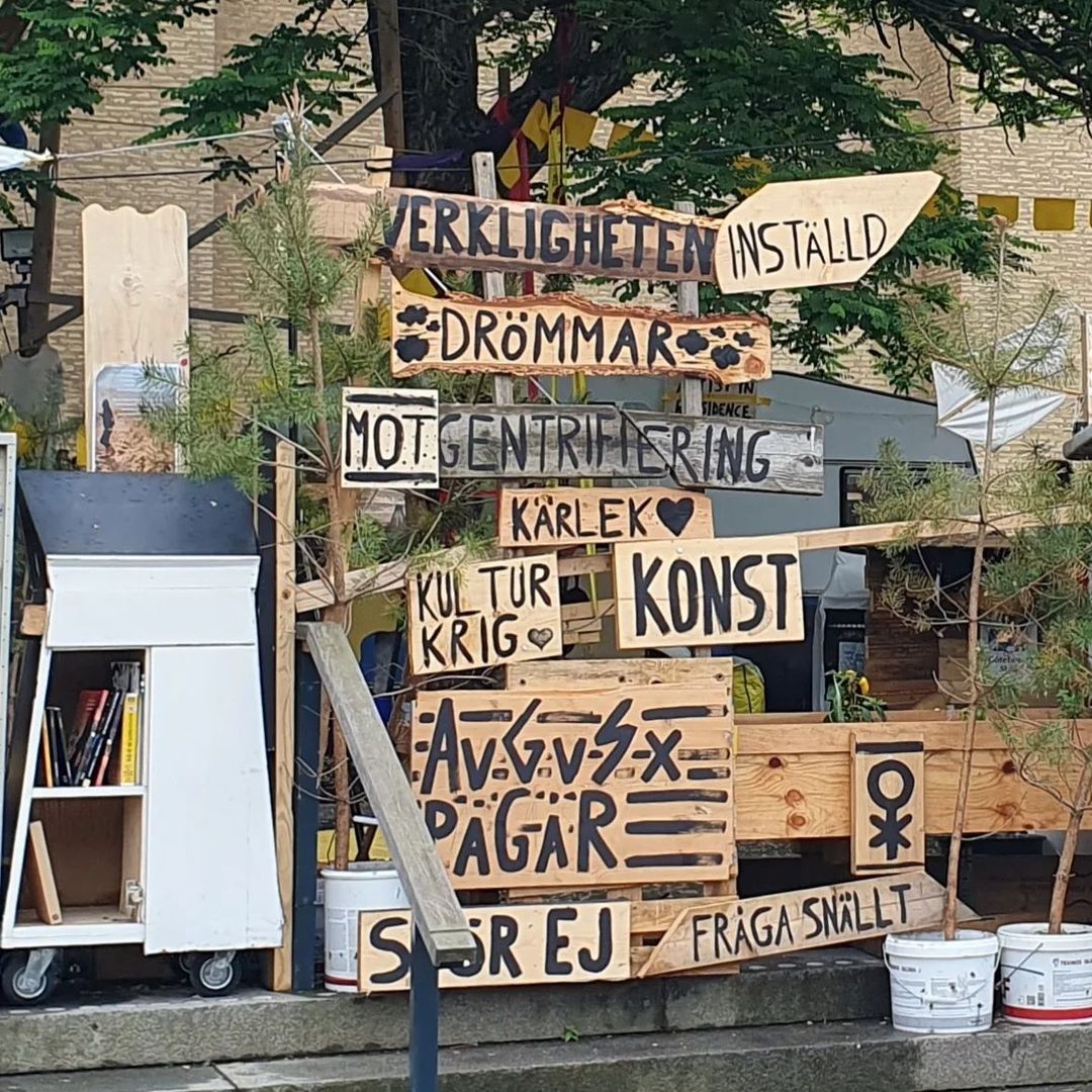 A rustic signpost of reclaimed wood with black painted text among plants. En vägvisare av återvunnet trä med svartmålad text bland små träd.