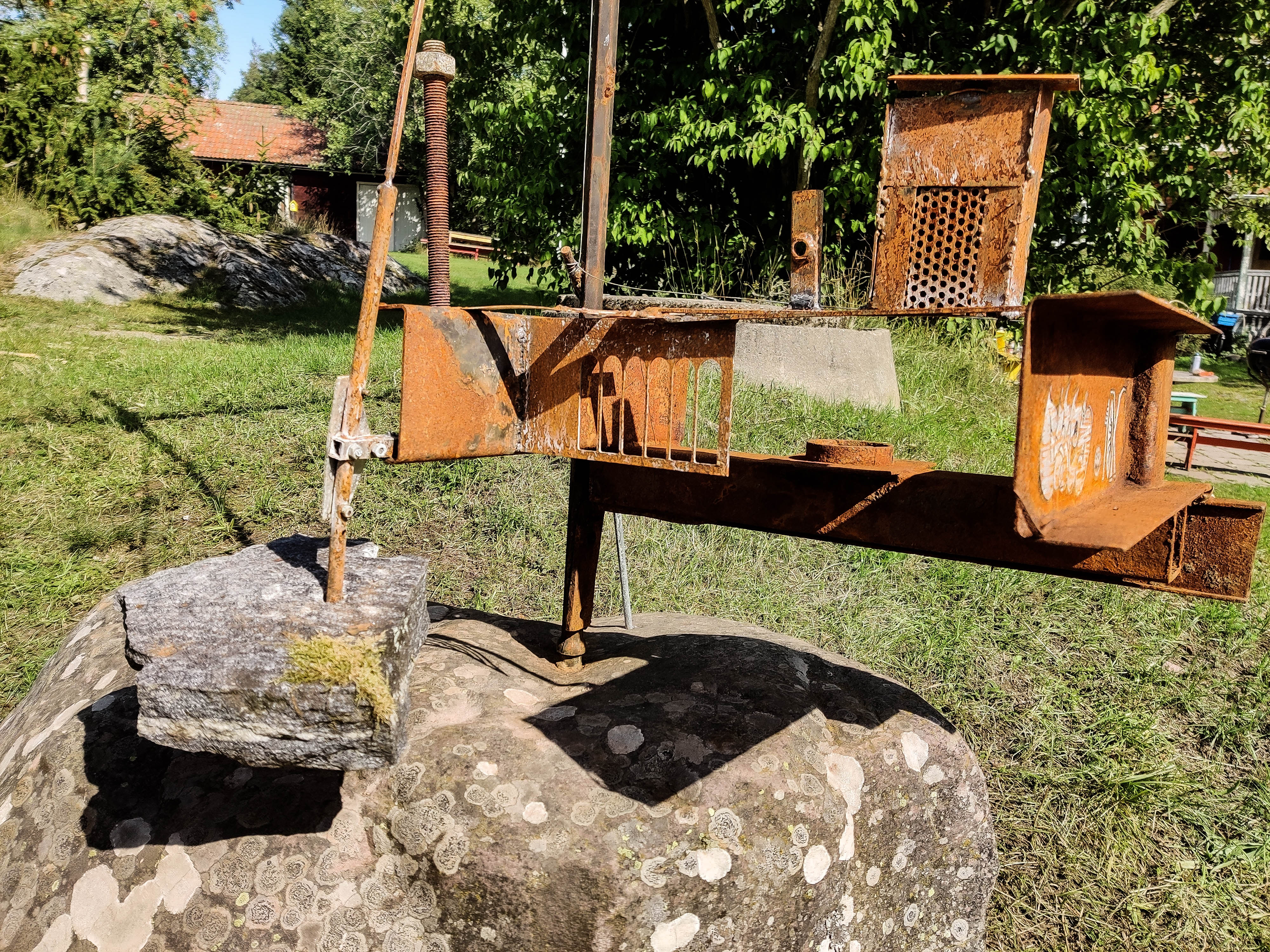 A rusted steel structure balancing on a large outdoor boulder. En rostig metallskulptur som balanserar på en stor sten utomhus.