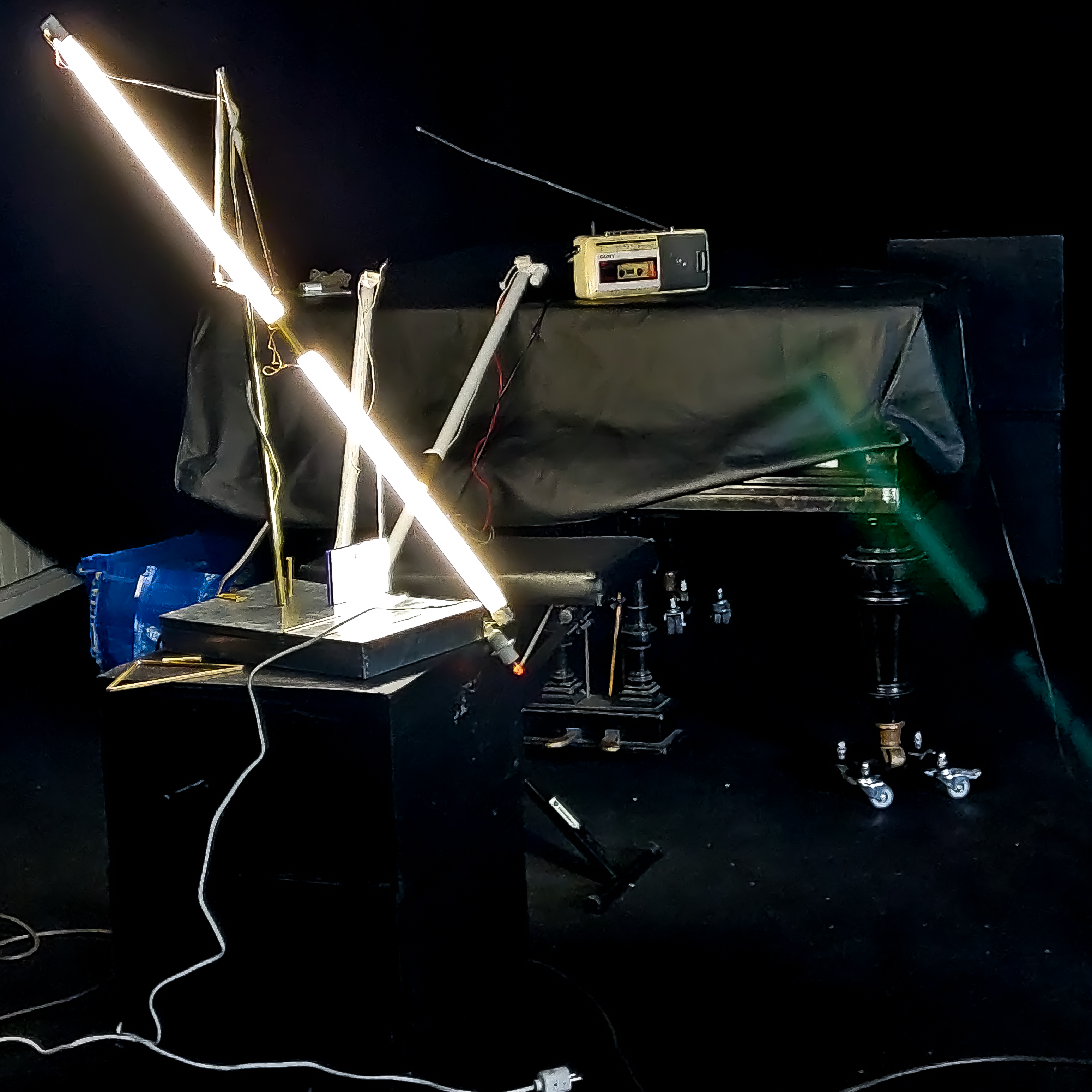 A bright fluorescent tube on a black stand, a boombox, and a tarp-covered piano. Ett ljust lysrör, en radio och ett täckt piano.