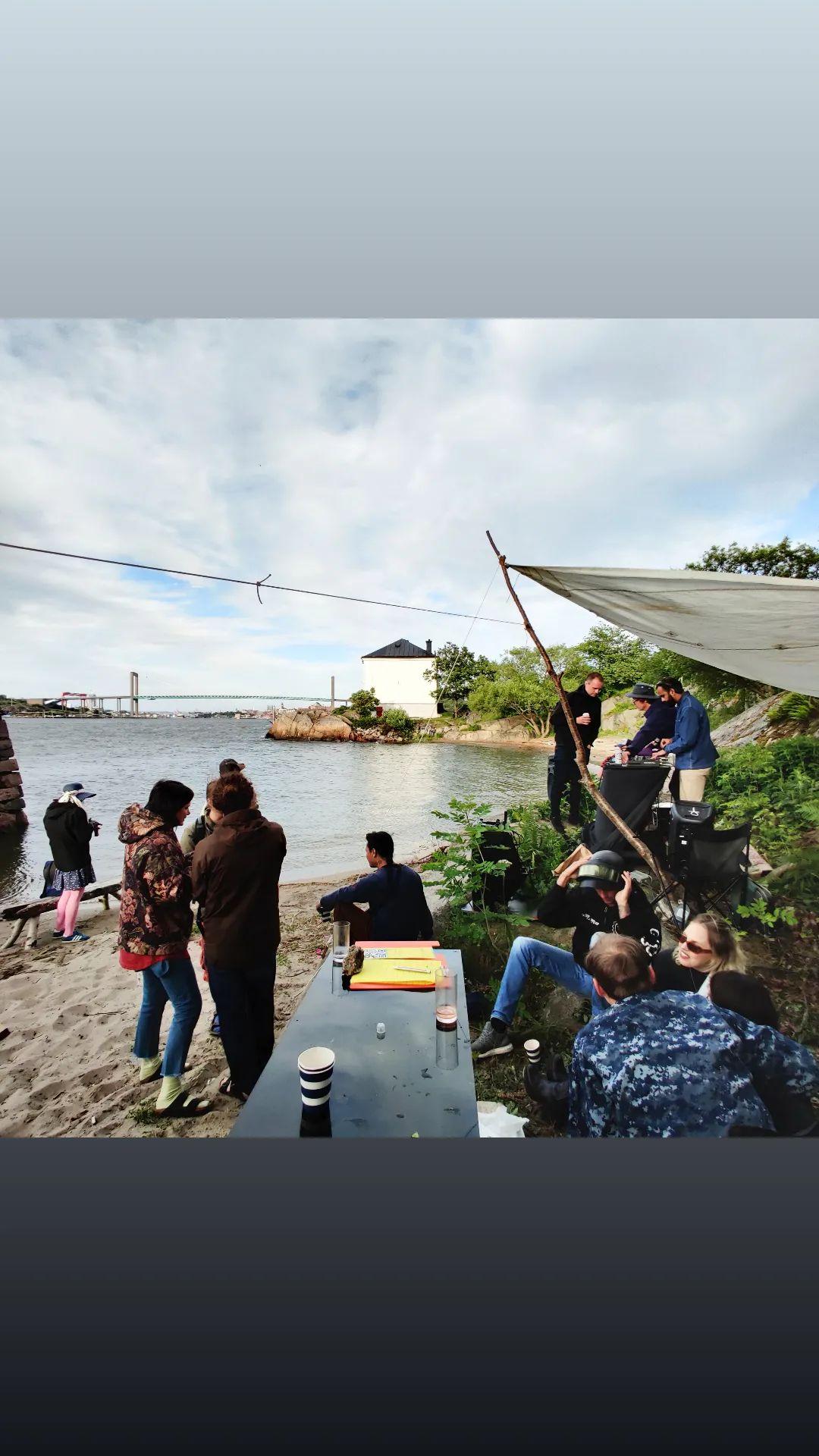 Group gathered by a DJ setup under a canvas tarp on a beach. Grupp samlad runt DJ-utrustning under en duk på en strand.