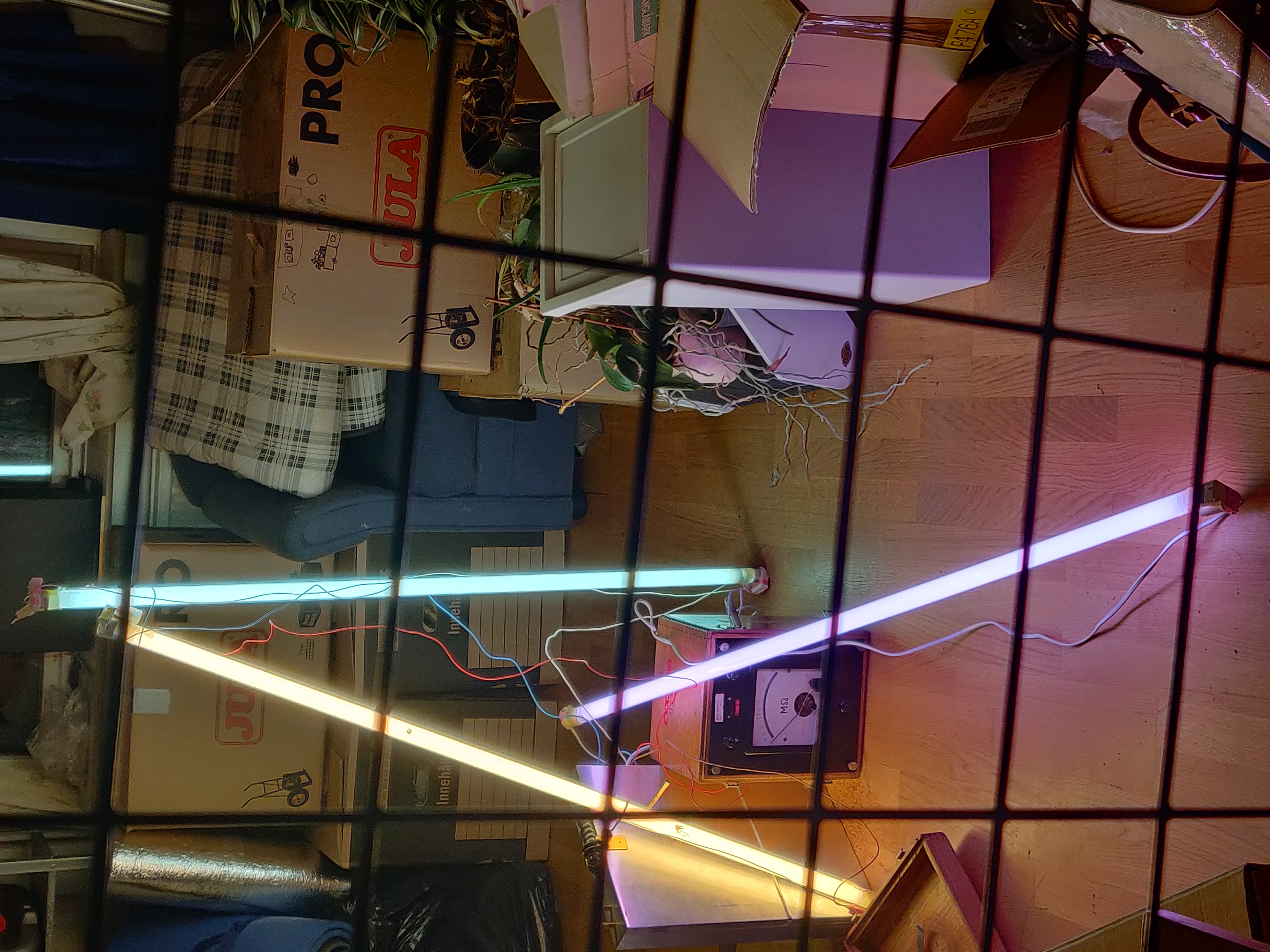 Three glowing light tubes lean on vintage electronics behind a grid. Tre lysande rör lutar mot äldre elektronik bakom ett metallnät.