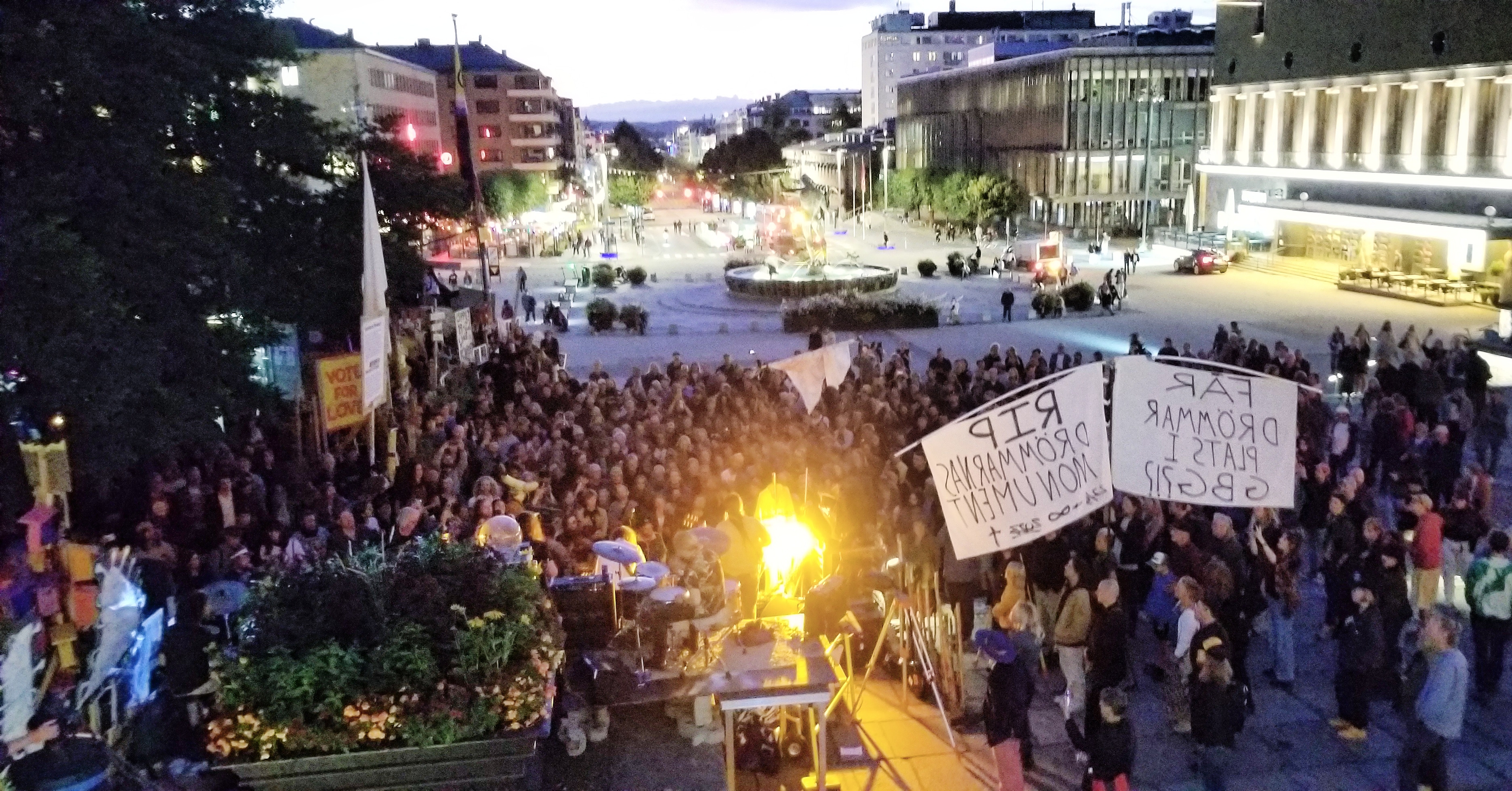 Large crowd gathers around a bright stage with fabric protest banners. Stor publik vid en upplyst scen med protestbanderoller i tyg.