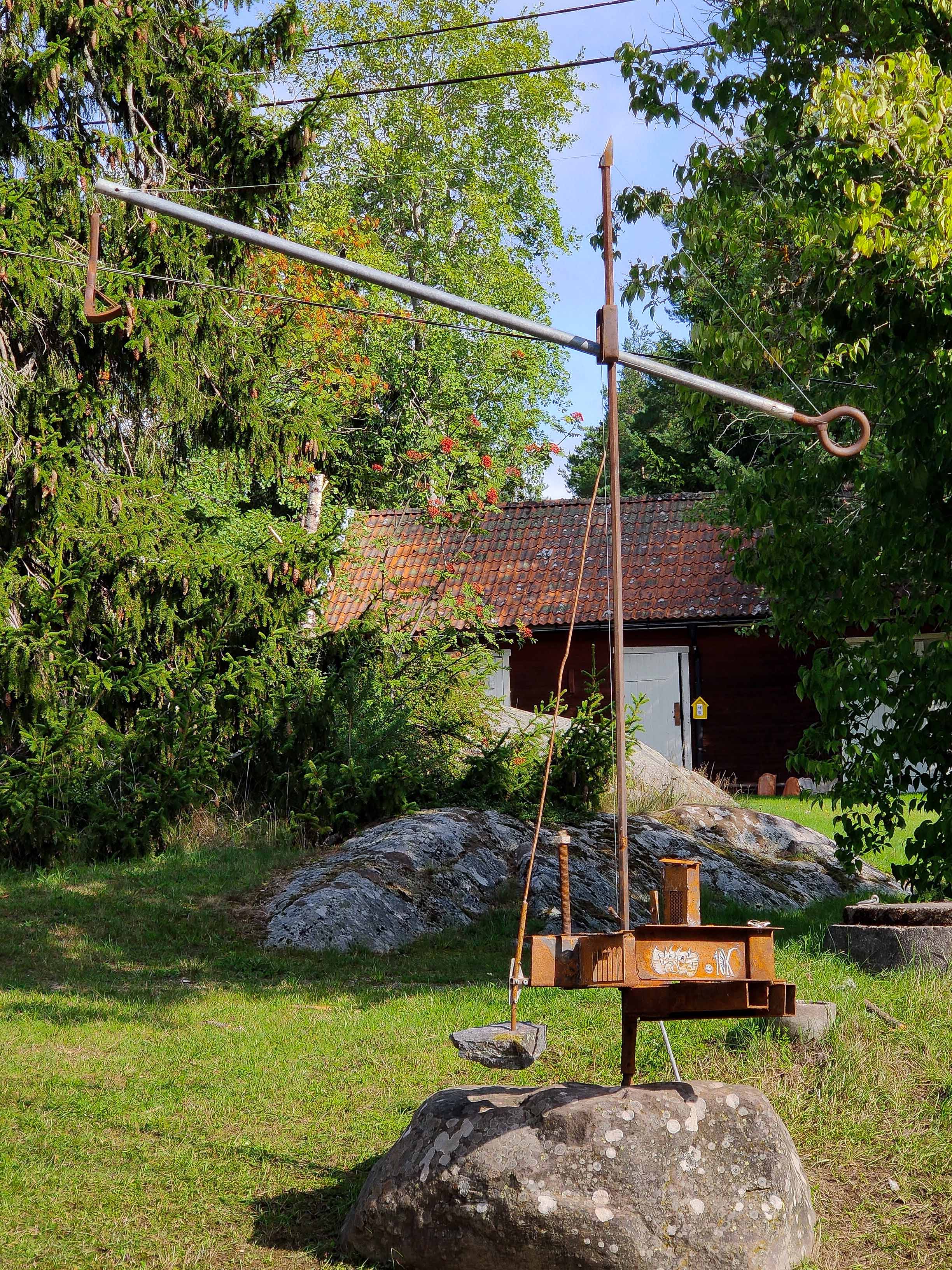 Rusty steel lever sculpture balancing a hanging rock, set outdoors. Rostig stålskulptur som balanserar en hängande sten utomhus.