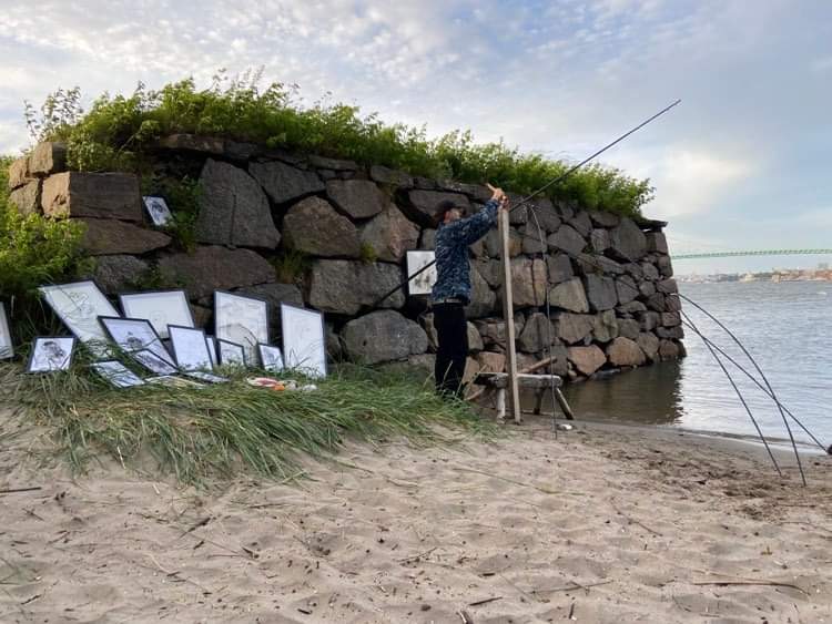 Artist on a beach setting up long metal rods near framed drawings by a stone wall. Konstnär på en strand sätter upp metallstavar nära inramade teckningar.